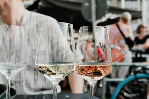 Ein Glas Weißwein und ein Glas Rosé auf dem Tisch, im Hintergrund Gäste – Weinprobe zu Hause planen